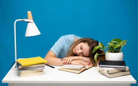 Tired student sleeping at her desk with books while having a lot of work. Busy schedule in college, workplace, sleep deprivation concept.の写真素材