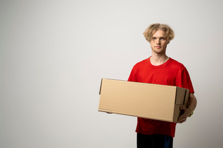 Delivery man in red uniform isolated on white background, studio portrait. Male employee working as courier dealer hold blank cardboard box. Service concept. Mock up copy space.の写真素材