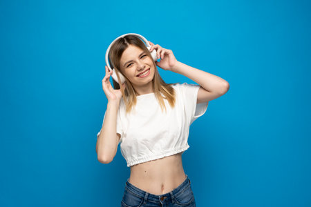 Young blonde woman in the white t-shirt with headphones listening music .Music teenager girl dancing against isolated blue background.の写真素材