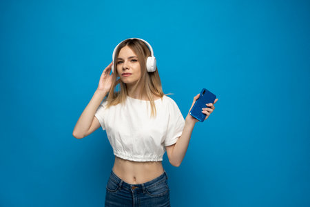 Young blonde woman in white t-shirt with headphones listening music. Music teenager girl dancing against isolated blue background.の写真素材