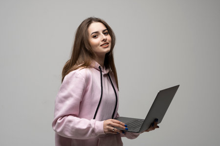 Attractive brunette woman 20s wearing pink hoodie standing working on laptop pc computer isolated on grey wall background studio portrait.の写真素材