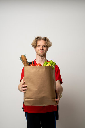 Delivery man guy employee man 20s wear red uniform work as dealer courier hold craft brown paper bag with grocery food vegetables isolated on white background studio.の写真素材