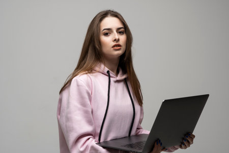 Attractive brunette woman 20s wearing pink hoodie standing working on laptop pc computer isolated on grey wall background studio portrait.の写真素材