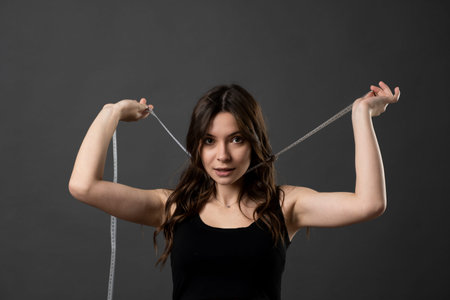 Young woman with slim body wear black t-shirt hold measure tape on neck look in a camera isolated on...