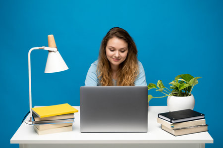 Focused woman working at desk with laptop, books, and plant on vibrant blue backgroundの写真素材