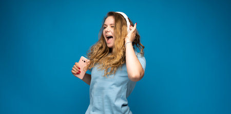 Happy young woman enjoying music in headphones with smartphone in hand on blue backdropの写真素材