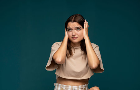 Young woman with brown hair covering ears with hands and uncertain expression