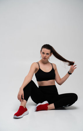 Young fitness woman in black sports bra and leggings sitting on a studio floor, pausing from workout with strong body language, focusing on wellness and healthy lifestyleの写真素材