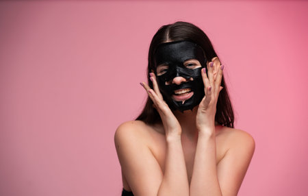 Woman enjoying a charcoal peel-off face mask, smiling while applying skincare product to her face during a beauty routine, standing against a pink studio backgroundの写真素材