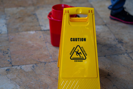 Bright yellow wet floor warning sign on tiled floor with red bucket in background, highlighting...
