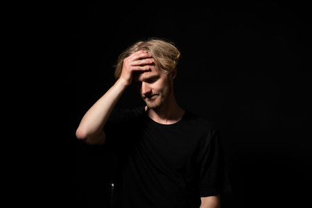 Young man experiencing headache or regret, holding hand to forehead, feeling pain, stress, or...