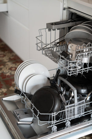 Dishwasher rack full of dirty dishes, plates, frying pan, and cutlery, ready for a wash cycle in a...