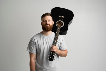 Bearded young man in grey t-shirt holding black acoustic guitar on shoulder