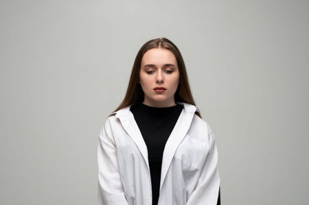 Young woman feeling sad and pensive, looking down with a thoughtful expression on a grey studio...