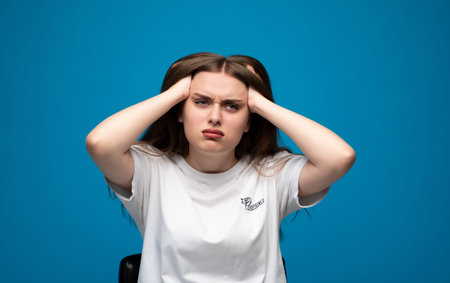 Stressed young woman enduring headache and pressure, showing anxiety and frustration on blue backgroundの写真素材