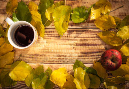 Top view autumn composition with yellow and green leaves laying on burnt wooden background with a cup of tea and apple. With free space for text on itの写真素材