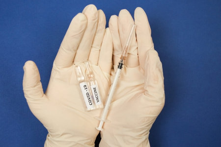 On a blue background, two vials of Covid-19 vaccine and a syringe lie in the hands of medical gloves