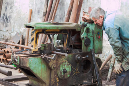 A worker saws a steel billet on an old oil-stained band sawの写真素材
