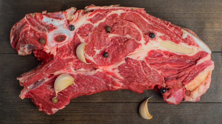 A piece of fresh red beef with cloves of garlic and peas of pepper lies on a dark cutting board....