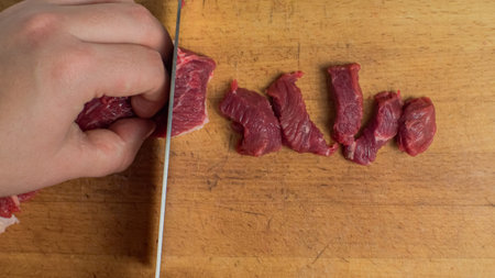 On a chopping board, a hand and a knife slice the red beef meat into chunks. Close-up