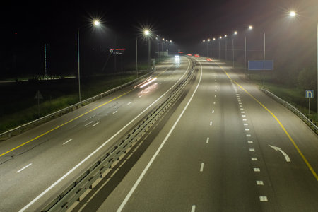 High-speed suburban highway in the light of streetlights. There are yellow and white markings on the asphalt. There are signs on the side of the road. Night scenesの写真素材