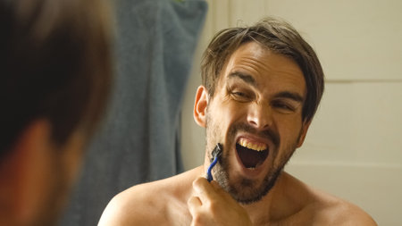 A shirtless man in front of a mirror, shaving with a blue razor without foam and screaming in pain. Close-upの写真素材