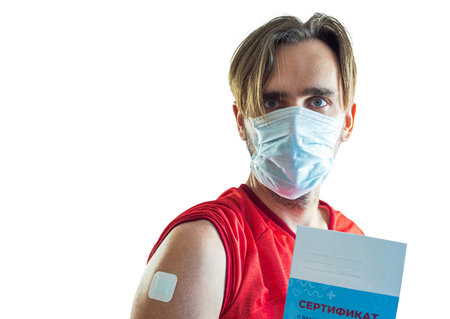 A man with a plaster pasted on his shoulder and a medical mask holds a vaccination certificate in his hand. Translation: certificate of vaccination against COVID-19のeditorial素材