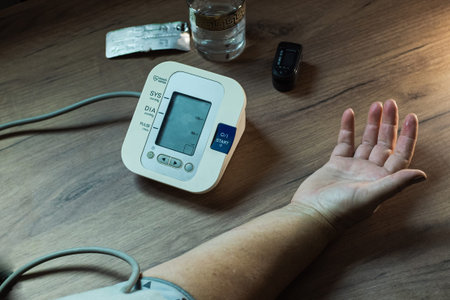 An elderly woman measures her blood pressure on an automatic tonometer. There is a glass of water,...