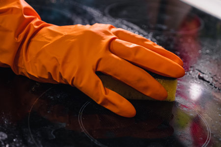 An orange glove with a yellow sponge cleans a dirty cooktop. Home Cleaning Concept