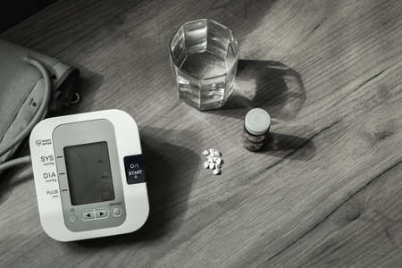 On a brown wooden table there is a glass of water, a bottle of pills, and a blood pressure monitor. A handful of pills are on the table. View from above. Black and white photo. Health Conceptの写真素材