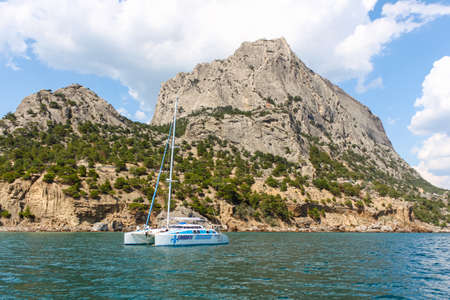 A yacht with a lowered sail against a rocky shore. There are white clouds in the sky. Republic of Crimea, Sudak, July 27, 2013のeditorial素材