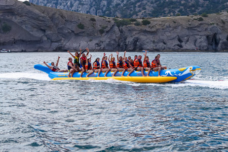 A crowd of people in life jackets in the sea on an inflatable banana against a rocky shore. Happy people at a seaside resort. Republic of Crimea, Sudak, July 27, 2013のeditorial素材