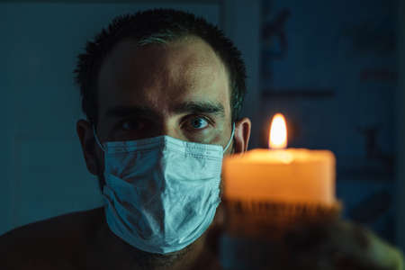 A close-up of the face of a white man wearing a medical mask against the background of a burning candle. The concept of grief in a pandemicの写真素材