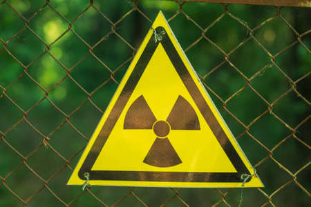 Close-up of a yellow triangular radiation sign on a wire mesh. In the background is a green forest. Warning of dangerの写真素材