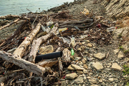 Logs, branches, plastic bottles and other debris are lying on the seashore. The stone beach is littered with garbage. The concept of ecologyの写真素材