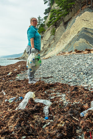 A woman volunteers with a bag full of garbage on a beach covered with plastic bottles. An...