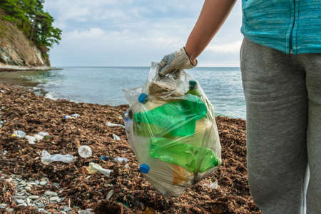 In the hand of the volunteer is a full bag of plastic bottles collected on the seashore. In the background is a polluted beach, the sea and the mountains. Eco-activist cleans the beach from pollutionの写真素材