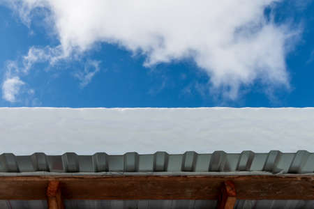 The roof of the metal profile shed covered with a thick layer of snow. In the background is a blue sky with clouds. A bright winter dayの写真素材