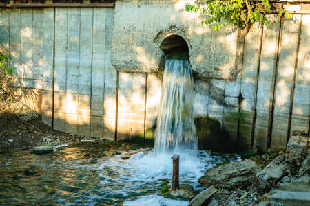 Water flows into the river after the sewage treatment plant through a pipe in the concrete wall. In the foreground is trash and concrete debris. The concept of ecologyの写真素材