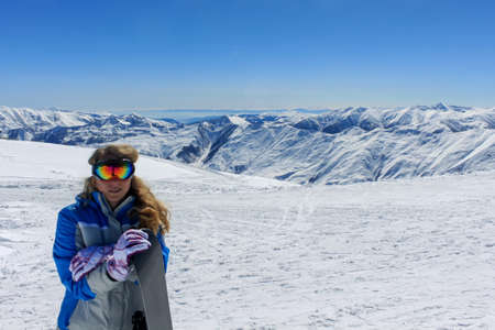 A snowboarder in ski goggles and loose hair is leaning on a board. In the background snowy mountains and clear blue sky. Active winter holidays in the mountainsの写真素材