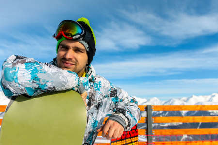 A Caucasian man in snowboarding clothes with a hat and goggles on his head, standing leaning on his snowboard. In the background is a wooden fence, snowy mountains and a blue sky with cloudsの写真素材
