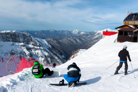 At the top of the ski slope, snowboarders strap on their boards and skiers are on their way. In the...