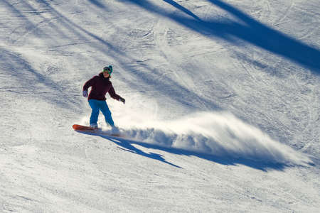 A woman is hurtling down a ski slope on a snowboard. A beautiful plume of snow flies out from under the board. There are long shadows on the slope. Active recreation during the winter vacationsの写真素材