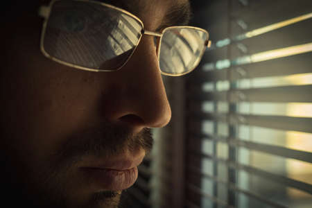 A Caucasian man with a beard and mustache and glasses stands at a window with blinds. There is a beautiful reflection of the blinds in the glasses. Close-upの写真素材
