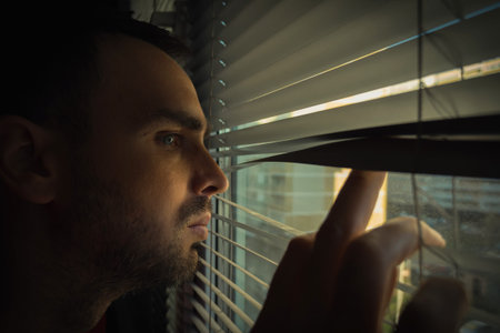 An unshaven Caucasian man looks out the window through the blinds. Close-up
