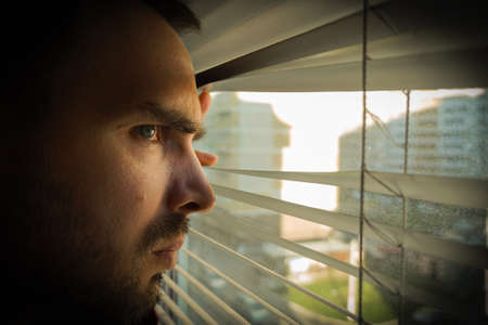 A man looks at the city through the dirty window through the blinds with a disgruntled look. Close-upの写真素材