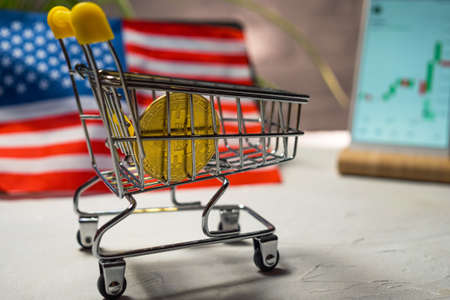 A shopping cart with Bitcoin in the background of the US flag and a smartphone with a graph of the BTC cryptocurrency exchange rate. blockchain technology. bitcoin in everyday lifeの写真素材