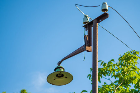 An old street lamp on a rusty pole with a broken bulb and torn wires against a background of green...