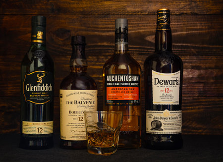 Bottles of 12 year old Balvenie, Auchentoshan, Glenfiddich, Dewars whiskey and a glass with ice on a dark wooden background. Premium brands of Scotch Whiskey. Russia, Krasnodar, December 11, 2021のeditorial素材