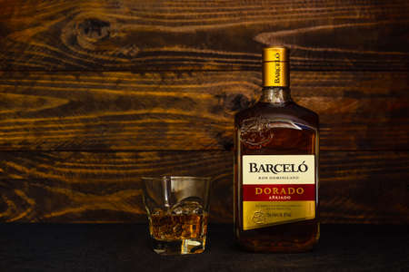 A bottle of Ron Barcelo Dominican rum, Dorado Anejado and a glass with ice on a wooden background. Ron Barcelo mild Caribbean rum is made from sugar cane juice. Russia, Krasnodar, December 11, 2021のeditorial素材